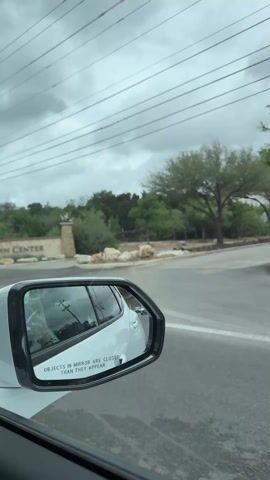 The video, shot from the inside of a moving car, shows the roadside as the vehicle passes by a "Town Center" sign. The sky is overcast and gray. Along the roadside, an orange traffic cone, concrete barriers, a red mesh construction fence, utility poles, and a shipping container are visible. The side mirror of the car is in the foreground.