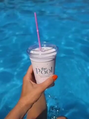 A hand with bright orange nail polish holds a plastic cup with a pink straw. The cup has a clear lid and contains a pinkish milkshake or smoothie. The cup has a label that says "the Pool" with smaller text below that is illegible. The hand and cup are submerged in bright blue water, likely a swimming pool, as evidenced by the ripples and the blurry legs of a person visible in the water below. The background shows a slightly out-of-focus view of a resort or pool area with buildings and some wooden structures. The lighting suggests it is a sunny day.