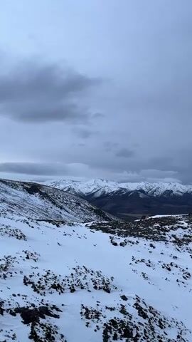 The video shows a snowy mountain landscape under a cloudy sky. The foreground is covered in snow with some dark patches of earth or rocks visible. In the middle ground, there is a steep snowy slope leading down into a valley. The background features a range of snow-capped mountains, with a darker, possibly wooded area visible between them. The sky is overcast with gray clouds. There are no people or animals visible in the video. The overall impression is of a cold, wintry, and remote environment.