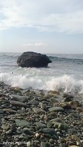The video opens to a low-angle shot of a rocky beach with waves crashing on the shore. A large boulder sits in the water, with waves breaking around it and sending white foam onto the beach. The sky is overcast with some scattered clouds. The camera pans slightly to the left, showing more of the waves and the rocks. The video ends with a large wave crashing against the boulder, creating a dramatic spray of water. The Infinix HOT 50 Pro logo appears in the bottom left corner of the screen.