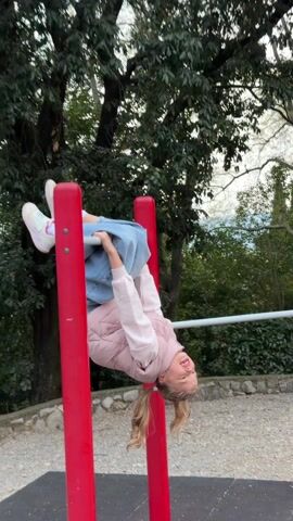 A young girl hangs upside down from a horizontal bar at a playground. She is wearing a pink puffy jacket, light blue jeans, and white sneakers. Her blonde hair is in pigtails. She smiles while looking at the camera. The bar is between two red poles. Trees are in the background.