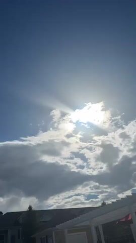 The video shows a sky with clouds. The sun is peeking through a large cloud, with rays of light shining through. There is a building with a white pergola structure in the lower right corner of the frame. The building appears to be a residential home or a clubhouse. The sky is a clear blue, and the clouds are white and gray. The sun is bright and appears to be setting or rising.