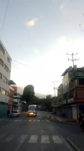 A street scene with buildings on either side. A white bus with red and pink stripes is driving down the middle of the street. There are white pedestrian crossing lines painted on the road. A few cars are parked on the side of the road. The sky is bright, suggesting it is daytime.