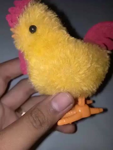 A hand holds a small, fluffy yellow toy chicken with a red felt comb and wattle, black bead eyes, and orange plastic beak and legs. The hand slowly rotates the toy, showcasing its front and side profiles against a plain, light-colored background.