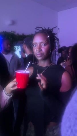 A woman with dreadlocks and a nose ring holds a red plastic cup with a straw in a dimly lit, crowded indoor setting. She wears a black sleeveless top and camo pants. Other people are visible in the background, some holding similar red cups. The lighting has a purple hue.