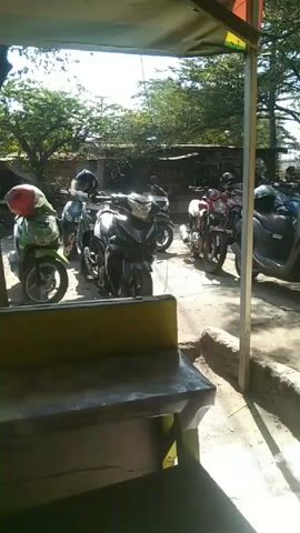 The video shows a row of parked motorcycles outside. The perspective appears to be from behind a counter or table looking out through a window or opening. The motorcycles are of various colors and styles, including what appear to be scooters and larger motorbikes. Some of the motorcycles have helmets on them. In the background, there are trees, buildings, and a clear sky. The lighting suggests it is daytime, likely sunny. There are no people clearly visible interacting with the motorcycles.