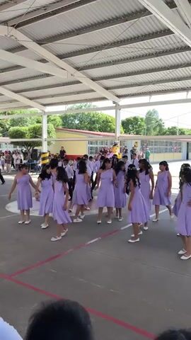 A group of young girls wearing matching lavender dresses are dancing in a formation outdoors under a covered structure. They are on a paved surface with red and white lines marking boundaries. Behind them, a building with yellow and red accents is visible, and other people, some appearing to be students and adults, are gathered around watching. The overall scene suggests an outdoor school event or performance.