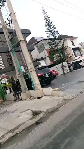 The video appears to be a handheld recording from inside a moving vehicle, likely a car, as evidenced by the blurred foreground and the perspective of looking out a window. The scene is a street in Moba, Nigeria.

The primary subjects are buildings lining the street, cars parked or driving, and a few individuals. We see several structures that appear to be commercial buildings, some with modern glass facades and others with more traditional designs. One prominent building has a large triangular roof and the text "MANTHEL HOLDINGS" is visible on it. Another building is painted green and has the text "Thrive Cafe" above its entrance. A white van and several dark-colored cars are present on the street and in front of the buildings.

There are at least two people visible in the video. In the first few seconds, two individuals are standing near the entrance of a building. Later in the video, a person in a striped top and dark pants walks across the scene from right to left. Another person is visible towards the end, also walking.

The activity appears to be the vehicle driving along a street, with the camera capturing the passing scenery. There are power lines and poles running along the street, indicating urban infrastructure. The ground appears to be paved but with some debris and puddles, suggesting it may have recently rained or that drainage is an issue.

The time of day is unclear, but the lighting suggests it is daytime, possibly overcast, as there are no strong shadows. The overall atmosphere seems ordinary and everyday, capturing a slice of street life in a Nigerian city.