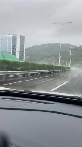 The video shows a view from inside a car driving on a wet road during a rainy day. The windshield has raindrops on it, and the windshield wipers are visible in the foreground. In the background, there are several tall buildings and a lush green hillside. The road appears to be a highway with multiple lanes, and there are streetlights and guardrails lining the side of the road. A white van is visible driving ahead on the road. The sky is overcast and grey.