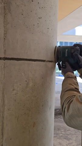 Here is a description of the video:

A construction worker uses a Makita concrete grinder on a gray concrete pillar, smoothing the surface near a horizontal crevice. The grinder, held with gloved hands, emits dust and noise as it moves across the concrete. The worker is wearing a tan jacket with the brand "Merrell" visible on the sleeve. The pillar is located outdoors under a partially covered area.