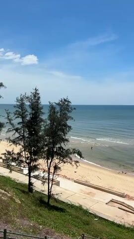 The video displays a wide view of a tranquil beach scene under a bright blue sky with a few scattered clouds. The camera pans slightly to the right, revealing a sandy beach with gentle waves lapping the shore. In the distance, a small dark boat is visible on the calm, blue-green ocean. To the left of the frame, two tall, slender trees stand on a grassy incline overlooking the beach. A concrete walkway runs parallel to the beach, with some construction materials or debris scattered nearby. The overall atmosphere is peaceful and serene, showcasing a typical coastal landscape on a sunny day.