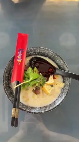 A bowl of soup with noodles, tofu, and greens is shown from an overhead perspective, along with a spoon and chopsticks. The chopsticks are in a red paper wrapper. The soup is in a dark grey patterned bowl on a light blue surface. The spoon is black and filled with a dark liquid, possibly a sauce. The tofu pieces appear golden brown. The greens appear to be fresh herbs. The chopsticks are resting across the rim of the bowl. The scene is well-lit with some reflections on the table surface.