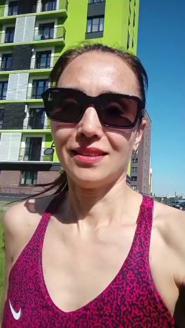 A woman with dark hair and sunglasses is walking outdoors. She is wearing a pink and black patterned tank top. Behind her is a tall, lime-green building with balconies. The sky is blue.