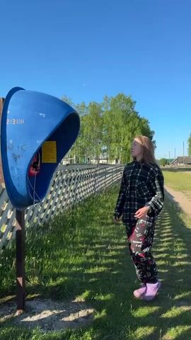 A young woman with long, light brown hair wears a black and white plaid shirt, black sweatpants with red pepper patterns, and purple slip-on shoes. She stands outdoors, facing a blue public telephone booth on a wooden post. The phone booth has a red payphone inside. The woman holds the phone to her ear with her right hand and uses her left hand to press a button on the phone. In the background, a white wooden fence runs along a dirt path, with green grass on both sides. Trees and a building are visible in the distance under a clear blue sky.