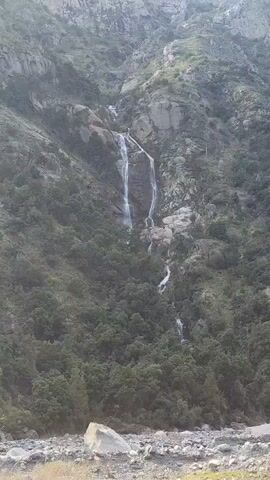 The video shows a waterfall cascading down a rocky, forested mountainside. The waterfall appears to be multiple streams of water flowing over ledges. The surrounding environment is lush with green trees covering the lower slopes of the mountain. The foreground shows a rocky riverbed, suggesting the waterfall feeds into a river. The lighting suggests it is daytime, but the sky is not visible. There are no people or animals visible in the video.