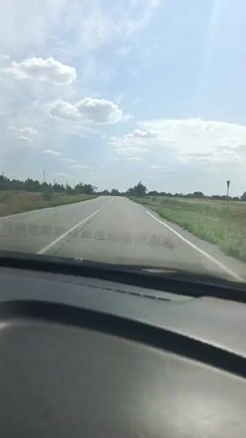 A dash cam view shows a paved road with white lane markings extending into the distance. The road curves slightly to the right. On either side of the road are grassy fields with scattered trees and shrubs. The sky is blue with scattered white clouds. The sun is visible, creating lens flare. The bottom of the video shows the interior of the vehicle, including the dashboard. The video is in landscape orientation.
