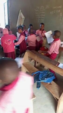 In an indoor setting resembling a classroom, a group of children are seen in a chaotic arrangement. They are dressed in pink shirts and dark maroon or red pants or skirts, possibly indicating a school uniform. The children are standing around wooden desks, some holding notebooks and papers. A few are smiling and appear to be animated or excited. The classroom has a window visible in the background, providing some natural light. Other classroom elements include a chalkboard on the wall and what looks like educational charts. Overall, the scene conveys a sense of a classroom environment with children who seem energetic and possibly engaged in an activity.