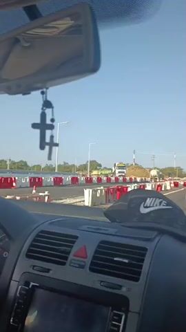 The video shows the interior of a car driving on a road. A cross hangs from the rearview mirror. In the foreground, a Nike hat is visible on the dashboard. The road is undergoing construction, with temporary barriers and traffic cones in place. Other vehicles, including a truck and a minivan, are visible on the road ahead. The sky is clear and blue, and trees and vegetation line the sides of the road.