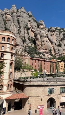 The video shows a wide shot of Montserrat Monastery in Catalonia, Spain. The monastery complex is built into a mountainside, with its buildings constructed from light-colored stone and featuring a mix of architectural styles. The buildings include a multistory tower, and some have tiled roofs. A large, paved plaza is visible in front of the buildings, with people walking around and some sitting on benches. The backdrop to the monastery is a rugged mountain range with tall, rocky peaks. The sky is clear and blue with only a few clouds.