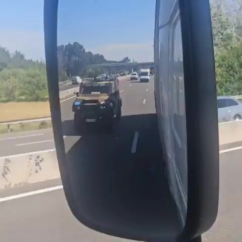 The video shows a vehicle driving on a highway, as seen from the side mirror of a larger vehicle, likely a truck or bus. Other cars and vans are also traveling on the highway. A green and brown armored vehicle is seen in the side mirror as well. The sky is clear and blue. The road is a light grey asphalt.  Green foliage is seen along the side of the highway. A truck with a blue trailer overtakes the filming vehicle. The traffic appears moderate.