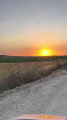 A steady outdoor shot captured at sunset shows a dirt road in the foreground that leads into a field on the right. Another field of grass and trees is visible on the left, which extends into the distance. The sky is clear, with shades of blue fading into orange and yellow as the sun sets. The sun is visible as a bright yellow circle in the middle of the frame. The video also captures the top of a vehicle in the bottom right corner, with a single amber light on its dashboard glowing brightly.