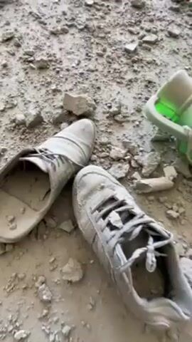 The video shows the interior of a room that has been heavily damaged, likely by an explosion. The floor is covered in debris, including pieces of concrete, broken glass, and personal items. A pair of shoes lies in the foreground. Three windows are visible in the background, with debris scattered near them and damage to the surrounding walls. Clothing is partially hanging out of one window. The walls are heavily damaged, with large chunks of concrete missing. There are several ceramic vessels on the floor, some intact and some broken. The overall impression is one of devastation and destruction.
