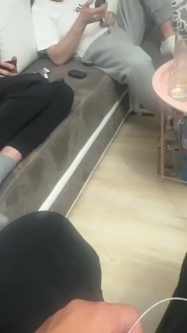 Here's a description of the scene:

A group of young men are relaxing indoors, likely at a home or apartment. One man is lounging on a gray couch wearing a white t-shirt and gray sweatpants. He is using his phone. Another person is visible with just their legs in black pants. A small, round, pink side table stands nearby with several glasses of orange juice. Other bottles of water and juice are visible under the table. The floor is light wood laminate. Music is playing in the background.