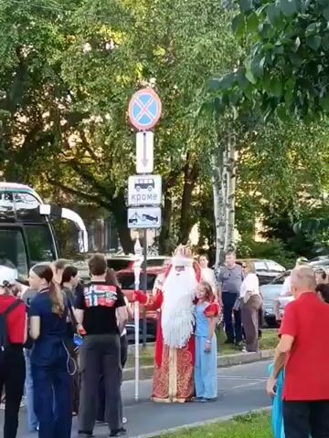 A group of people are gathered on a sidewalk near a street. In the center of the group is a person dressed as Santa Claus, wearing a red and gold robe, a white beard and hat, and holding a staff. A young girl in a blue outfit stands next to Santa Claus. Behind them are several road signs, including a circular sign with a blue background and a red X, and a rectangular sign with text and images. A white bus is visible on the left side of the frame. Other people, some carrying backpacks, are standing around. The environment appears to be outdoors with trees and parked cars in the background. The lighting suggests it is daytime, likely late afternoon due to the long shadows.