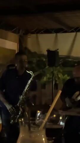 This video captures a live music performance by a band in what appears to be a bar or restaurant setting. Two male musicians are prominently featured. On the left, a man wearing a blue shirt and glasses plays a saxophone, actively engaged in his performance. To his right, another man, also wearing a dark shirt and glasses, plays an electric guitar. A third individual is partially visible in the background, suggesting more band members.

The setting is dimly lit, indicating an evening or nighttime event. Dynamic stage lighting changes from a warm red glow to a cooler green, illuminating the musicians and the performance area. In the foreground, several drinks are visible, including cocktails in glasses with straws and what appears to be a glass of beer, suggesting an audience enjoying the music. The background features a simple backdrop, possibly a painted scene or mural, and some artificial foliage, adding to the relaxed ambiance. A speaker on a stand is positioned behind the musicians. The overall atmosphere seems lively and entertaining.