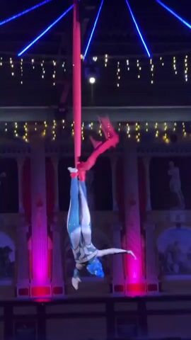 A solo aerialist performs a dance routine suspended by a single red silk fabric. The aerialist, wearing a blue and white patterned costume, is positioned upside down, with their legs wrapped around the fabric. The background features a theatrical set designed to resemble a Roman or Greek-style building with columns and murals. Pink and green stage lights illuminate the scene, casting vibrant colors on the backdrop and the performer. Above, strings of yellow lights and blue neon strips create a festive atmosphere. The setting appears to be an indoor performance space, possibly a circus tent or theater, during an evening show. The aerialist's movements are graceful and fluid, showcasing strength and flexibility as they manipulate the fabric. The overall mood is one of spectacle and artistic expression. There is no visible text in the media file.