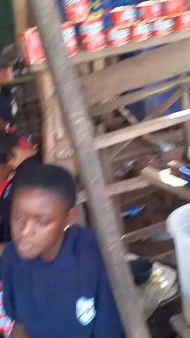 The video shows a young person scooping rice from a large container onto a woven mat at what appears to be an outdoor market or store. The surrounding area looks cluttered, with buckets, baskets, bags, and wooden structures scattered around. Other stalls and people are visible in the background. The young person is wearing a white tank top.