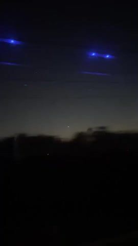 This media file captures a nighttime scene from a moving vehicle, likely a bus, given the presence of blue LED lights lining the ceiling. The view is through a window, with the landscape outside blurred due to motion. The setting appears to be rural or suburban, with a dark silhouette of trees and possibly hills against a dusky sky.

The sky exhibits the soft gradient of twilight, transitioning from a deep blue overhead to hues of orange and yellow near the horizon, suggesting the time of day is either dawn or dusk. Several thin, dark lines stretch horizontally across the frame, which could be power lines or other overhead infrastructure. Small, faint lights are visible in the sky, possibly distant stars or even planets.

The foreground is dominated by a dense, dark mass representing the landscape passing by. The motion blur emphasizes the sense of travel and movement. The blue lights on the ceiling of the vehicle are the most prominent artificial elements, casting a cool glow. There is no visible text in the media file. The overall mood is serene and slightly mysterious, evoking a sense of peaceful journey through the fading light. The location context of Hod HaSharon, Israel, places this scene within a specific geographical area.