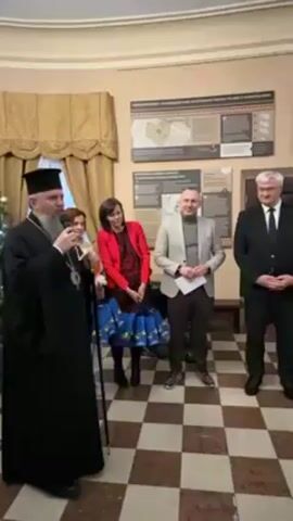 A man in a black clerical robe is speaking into a microphone. He is standing in a room with four other people. To his left is a Christmas tree. The others are a woman in a red jacket, a man in a grey jacket, and a man in a black suit. They are standing on a checkered floor. Behind them are framed images and text on the wall.
