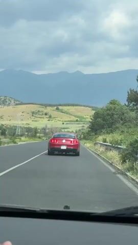 The video shows a red Ford Mustang driving on an asphalt road in a rural setting. The Mustang is the primary focus, seen from the rear, moving away from the viewer. It has dual exhaust pipes and a license plate that reads "PBTB 0538." The surrounding environment consists of rolling hills covered in sparse greenery, with a backdrop of mountains under a cloudy sky. A roadside guardrail and some foliage are visible on the right side of the road. The footage is shot from the perspective of someone inside another vehicle, looking through the windshield. The camera is relatively stable.
