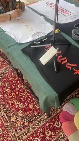 A person is painting a design on a white t-shirt with gold paint.  There are other t-shirts on the table, one white and one black, both with painted designs. The person is wearing bracelets and holding a small jar of gold paint and a thin brush.  The table is covered with a green cloth, and a red patterned rug is visible underneath.  A microphone stand is positioned near the t-shirts.