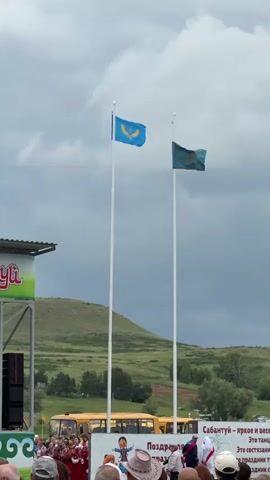 The video shows two flags flying on flagpoles against a cloudy sky. The flag on the left is blue with a yellow emblem. The flag on the right is green with a yellow emblem. There is a stage on the left side of the frame with speakers and a banner. In the background, there are green hills and a yellow bus. A crowd of people is gathered in front of a banner that says "ПОЗДРАВЛЯЕМ С ПРАЗДНИКОМ" which translates to "CONGRATULATIONS ON THE HOLIDAY" and "Сабантуй - яркое и веселое". Sabantuy is a Tatar festival. People in the foreground are mostly looking towards the stage or the flags. The overall scene appears to be an outdoor festival or event.