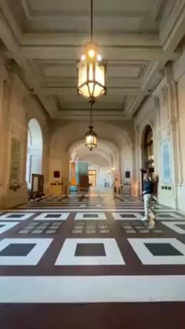 The video shows the interior of a grand building, possibly a museum or government building. The camera moves through a long hallway with a checkered floor of white, black and dark red squares. The walls are off-white with arched openings on both sides. Large, ornate light fixtures hang from the ceiling. A man in a blue jacket and khaki pants walks down the hallway. At the end of the hallway, a large arched iron gate stands open, revealing a view of the outside. People are standing near the gate, some entering or exiting the building. A crowd can be seen outside the gate in the distance.