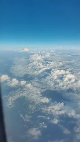The video shows an aerial view of clouds and the sky from an airplane. The perspective is looking down at a vast expanse of white and gray clouds, with patches of blue sky visible in between. The clouds appear fluffy and textured, with some areas brightly lit by the sun and others casting shadows. The horizon is visible in the distance, where the clouds meet the clear blue sky. The overall scene is serene and expansive, conveying a sense of being high above the Earth.