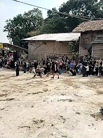 A crowd of people is gathered around a sandy or dirt area where performers are engaged in an activity. In the center, a performer in a costume resembling an animal, possibly a horse, is dancing or moving energetically. Other performers in black clothing are also present, some holding what appear to be props like whips or ropes. Spectators are observing the performance from the sides, some standing and others seated or crouching. The background shows buildings with corrugated roofs and bamboo structures, suggesting an outdoor event in a rural or semi-rural setting. The overall atmosphere seems to be a festive gathering or cultural performance.