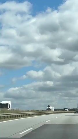 A shot from a vehicle traveling on a highway. The sky is partly cloudy with scattered patches of blue. Road signs are visible on the right side of the highway. Several vehicles, including cars and trucks, are on the highway. A bird is seen flying in the distance. The road has lane markings. The surrounding landscape appears to be rural with trees and vegetation.