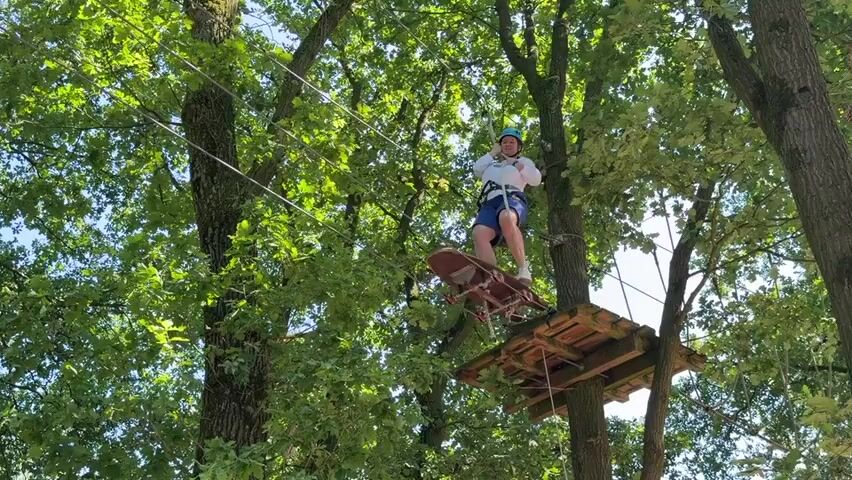A person wearing a helmet and a white shirt with blue shorts is riding a skateboard-like apparatus on a zip line through a forest. The person is suspended by a cable above the ground. The skateboard is suspended by cables attached to the person's harness. The background is filled with lush green trees and dappled sunlight. To the right, there is a wooden platform attached to a tree. Two other people are visible on the platform, looking at the person on the zip line. The scene appears to be a ropes course or adventure park.