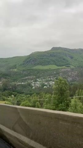 The media file captures a scenic view of a valley with a village nestled amidst rolling green hills and a prominent mountain range under a cloudy sky. The foreground is dominated by a concrete barrier, suggesting the footage is from a vehicle moving along a road. Beyond the barrier, a dense forest of trees blankets the lower slopes of the hills, giving way to scattered houses and buildings that form a small village in the valley. The village appears to have a mix of residential buildings and possibly some public structures, indicated by rooftops of various colors and sizes.

The scene is set in a rural mountainous landscape, likely in Skare, Norway, given the context. The time of day appears to be daytime, with diffused light due to the overcast weather. The clouds are heavy and grey, contributing to a muted, cool-toned atmosphere. There are no visible people in the frame. The activity taking place is simply the observation of the landscape from a moving vehicle. The prominent features are the natural elements: the lush green vegetation covering the hills, the imposing mountain in the background, and the sky filled with clouds. The concrete barrier in the foreground is a man-made object, serving as a visual anchor and indicating the perspective of the viewer.

Notable details include the texture of the forest, the rocky outcrops visible on the mountainside, and the neat arrangement of houses in the village. The overall impression is one of tranquility and natural beauty, typical of a mountainous region. The weather suggests a possibly cool or temperate climate. No visible text is present in the media file.