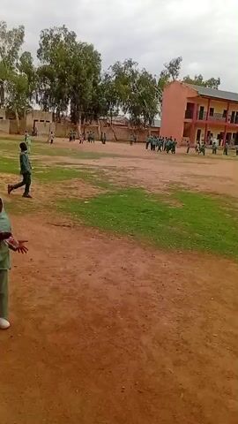 The video shows an outdoor scene, possibly a schoolyard, with several children engaged in various activities. In the foreground, a young child wearing a green headscarf and matching attire is walking or running towards the left of the frame, holding a plastic bottle. Their gait suggests movement and purpose. In the background, a group of children, mostly in similar green uniforms, are spread across a field. Some appear to be playing or gathering near a multi-story building that resembles a school. Other buildings are visible, some with solar panels on their roofs. The ground is a mix of dirt and patches of grass. Trees and vegetation dot the perimeter of the field. The sky is overcast, suggesting a cloudy day. The overall impression is of a school or community gathering on a relatively calm day with children actively participating in outdoor activities.