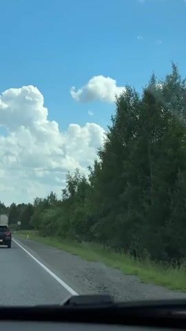 This is a vertical, first-person perspective video of a person driving on a highway. The sky is blue with white clouds, and there are trees on the right side of the road. There are several cars on the highway, including a black SUV in front of the camera and a white truck to the left. The car in front is moving, and the camera vehicle is keeping pace with it.