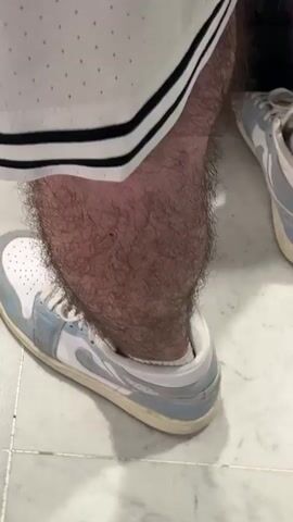 A close-up, low-angle shot shows a person's lower legs and feet, likely at Orly, France, suggested by the context of an airport location. The person is wearing white basketball shorts with black stripes along the side seams. Their legs are visibly hairy. On their feet are light blue and white low-top sneakers. A black rolling suitcase is partially visible behind the person's right leg. The floor is made of light-colored, possibly marble or ceramic, square tiles. The person appears to be standing still.