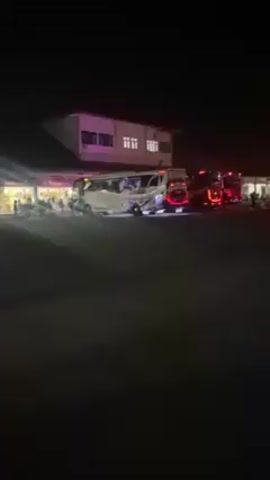 The video shows a bus terminal at night. Several buses are parked in a line, with their red tail lights visible. There is a building with windows in the background, and people are milling about near the buses. A light post shines brightly, casting a beam of light across the scene. The ground appears to be asphalt.