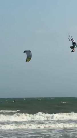 A kitesurfer in mid-air is captured in the frame, soaring above a choppy ocean. The kitesurfer, wearing a white shirt and blue shorts, is suspended by a kite connected by lines to their harness. Their surfboard is beneath them, and their body is angled as if performing a trick. In the background, another kite is visible, also in the sky, and a person is on a surfboard in the water. The sea is a deep blue, with white waves crashing on the shore in the foreground. The sky is a pale, hazy blue, suggesting a bright but possibly overcast day. The overall scene depicts a dynamic moment of extreme water sports.