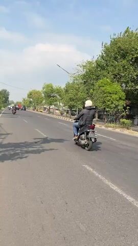 This media file captures a sunny day in Siring, Indonesia. The scene unfolds on a paved road lined with trees, indicating an outdoor setting, likely a street or highway. 

The primary subject is a person riding a black motorcycle. They are wearing a black jacket, blue jeans, and a white helmet, obscuring their face. The motorcycle itself is a prominent object in the foreground. In the background, several other vehicles, including motorcycles and cars, are visible on the road, suggesting moderate traffic. The surrounding environment is characterized by lush green trees and a clear blue sky with scattered clouds, pointing to a pleasant, possibly warm, time of day. The sunlight casts shadows on the road, further reinforcing that it is daytime.

The activity depicted is simply the act of riding a motorcycle. There are no overt signs of specific events or strong emotions. The overall atmosphere appears calm and ordinary, portraying a slice of everyday life in this Indonesian location. The visible text on the motorcycle's license plate reads "W 6333 YP".