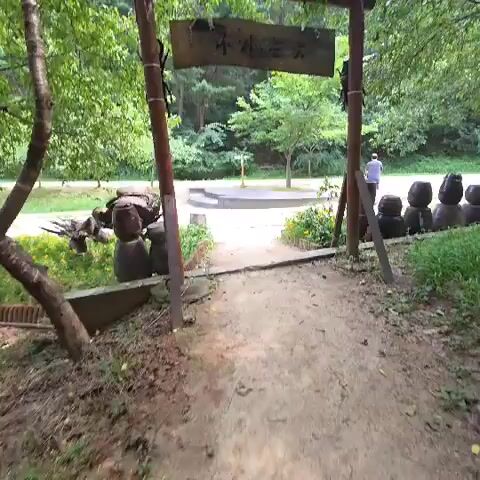 A first-person perspective video captures a path leading through a park-like setting. The path is made of dirt and gravel, bordered by grass and some decorative items. On the left side of the path, there are large ceramic jars and a metal sculpture of a bird. On the right side, there are several dark, rounded objects that resemble large stones or statues. The path leads towards a slightly elevated area with a curved, concrete structure, possibly a stage or seating area. In the background, there are trees and foliage, suggesting a natural environment. A person is visible walking away from the camera in the distance, near the curved structure. The overall atmosphere is peaceful and natural.