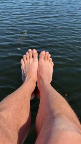 A low-angle close-up shows a person's bare feet dangling over the edge of a boat or dock, submerged in calm, dark blue water. The person's legs are visible above the ankles, with some hair on their shins. In the background, across the water, there's a lush green landscape with what appears to be lily pads or similar vegetation, and distant mountains under a clear blue sky with a few wispy clouds. The water ripples slightly. The camera slowly tilts upwards, revealing more of the scenic background.